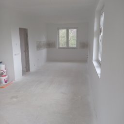 Gruszka usługi remontowo budowlane - Wnętrze pokoju w trakcie prac wykończeniowych; ściany i sufit pomalowane na biało, widoczne puszki z farbą, szpachlowane fragmenty ścian przygotowane pod malowanie, surowa podłoga.