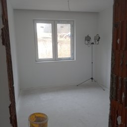 Gruszka usługi remontowo budowlane - Wnętrze w trakcie prac wykończeniowych: świeżo pomalowane na biało ściany, surowa podłoga, widoczne lampy na statywie i wiadro po farbie, fragment muru w surowym stanie.
