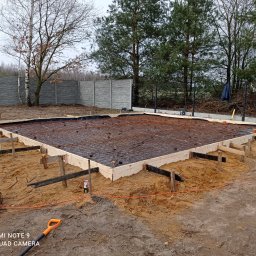 Gruszka usługi remontowo budowlane - Przygotowanie zbrojenia i szalunku pod fundament płyty betonowej na piaszczystym podłożu, widoczne siatki zbrojeniowe, drewniane deski szalunkowe, narzędzia budowlane i częściowo ogrodzenie...