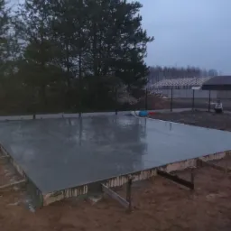 Świeżo wylana, mokra płyta fundamentowa z widocznym deskowaniem i wystającymi prętami zbrojeniowymi, na zewnątrz, w tle drzewa i częściowo ogrodzenie posesji.