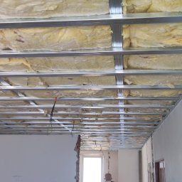 Gruszka usługi remontowo budowlane - Sufit podwieszany z wełną mineralną w trakcie montażu, widoczne profile stalowe i przewody elektryczne, w tle surowa ściana z widocznymi śladami po demontażu.