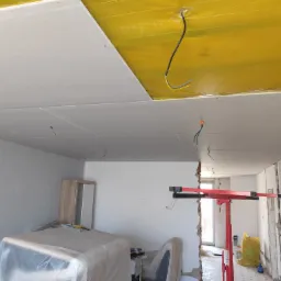 Wnętrze w trakcie montażu sufitu podwieszanego z płyt kartonowo-gipsowych; widoczne profile, żółta folia paroizolacyjna, podnośnik do płyt oraz wystające przewody elektryczne.