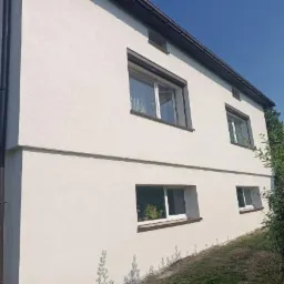 Dwukondygnacyjny budynek z jasną elewacją o fakturze baranka, brązowe ramy okienne i rynny, widok z dołu w słoneczny dzień.