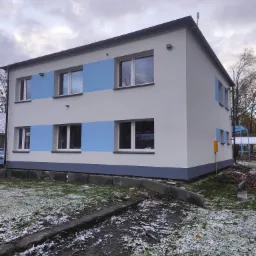 Dwukondygnacyjny budynek po renowacji elewacji z pomalowanymi na niebiesko pasami między oknami, widoczny z zewnątrz, z fragmentami śniegu na trawie.