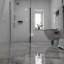 Nowoczesna łazienka z białymi płytkami, kabiną prysznicową z czarnymi ramami, wiszącą toaletą i rośliną w oknie, widok z niskiego kąta.