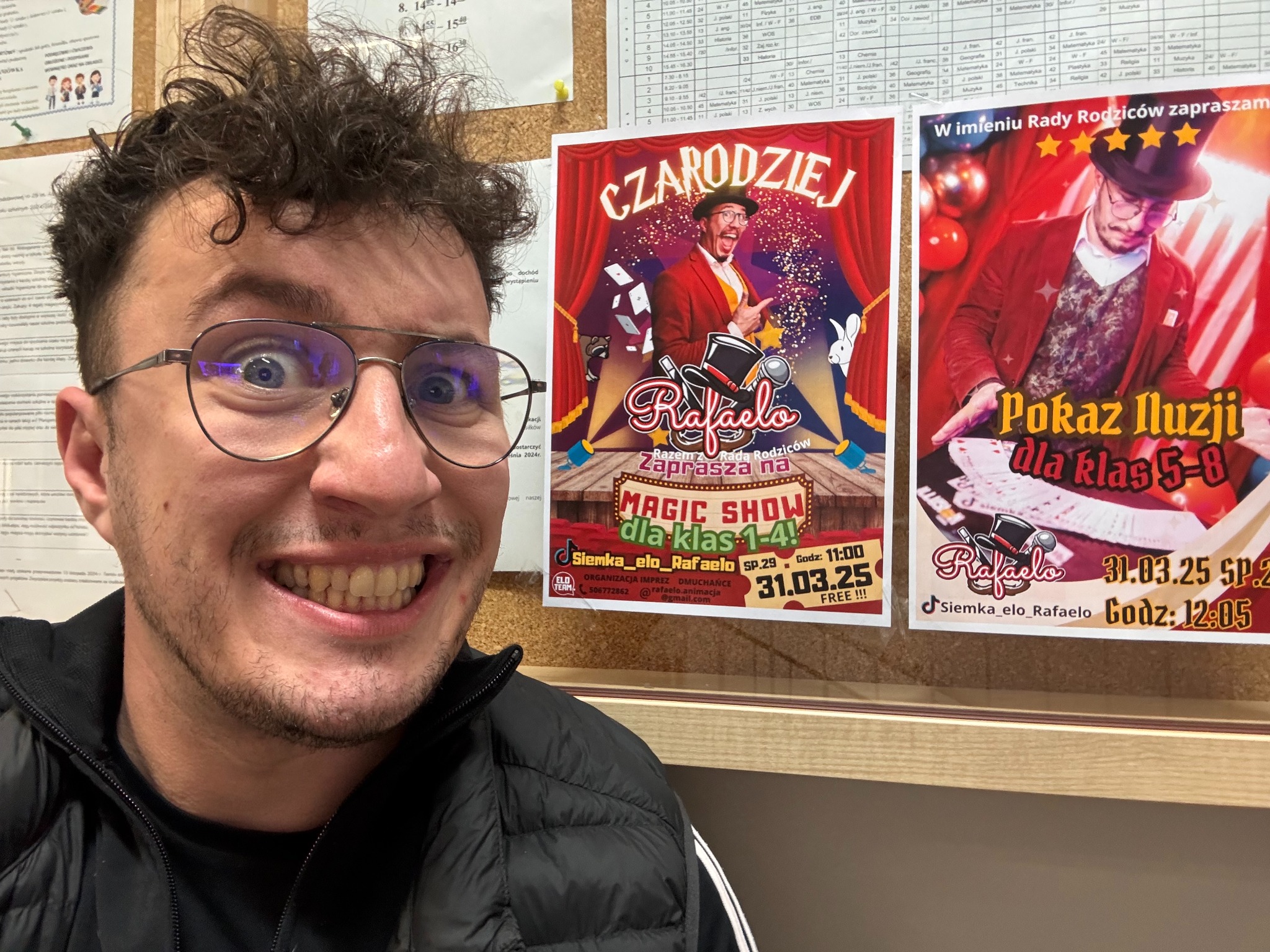 Uśmiechnięty mężczyzna w okularach robi selfie na tle plakatu z iluzjonistą Rafałem, zapraszającym na magiczne show dla klas 1-4, 31.03.25. Darmowe wejście.