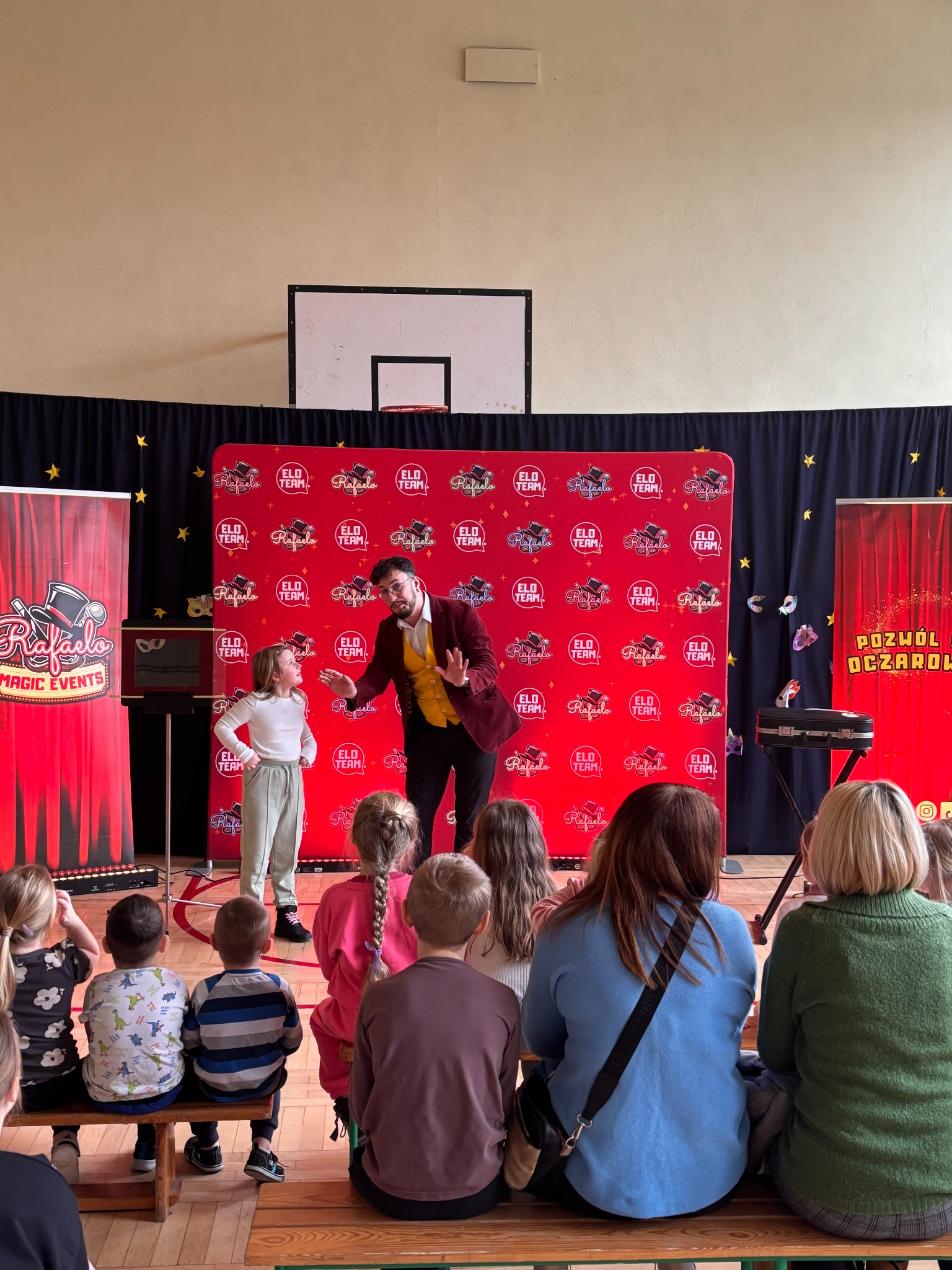 Iluzjonista w bordowej marynarce i żółtej kamizelce prezentuje trik dziewczynce na tle czerwonej ścianki z logo. Widzowie siedzą na ławkach.