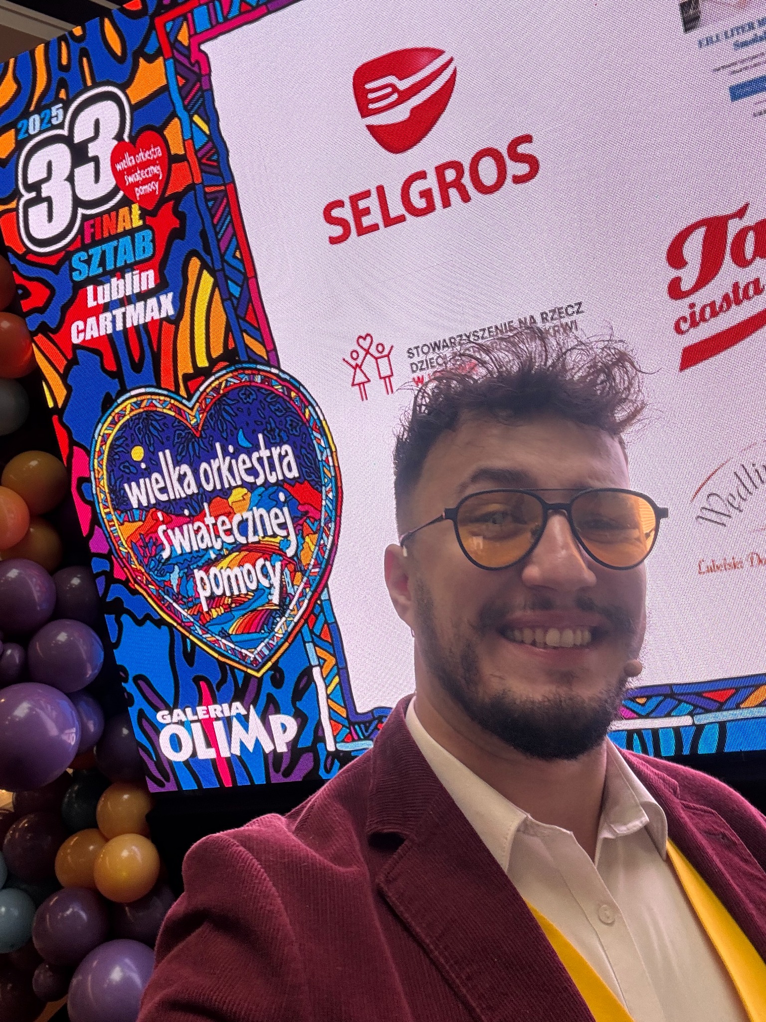 Uśmiechnięty mężczyzna w okularach na tle ekranu z logo WOŚP, Selgros i dekoracji balonowych. Wydarzenie charytatywne, finał WOŚP w Lublinie, Galeria Olimp.