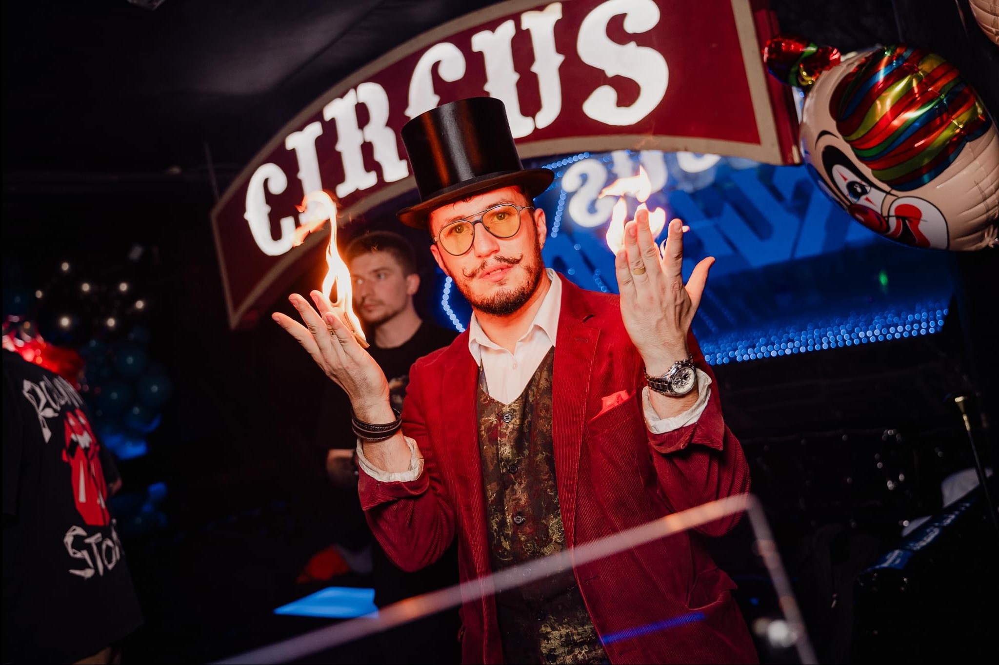 Iluzjonista w czerwonym fraku i cylindrze prezentuje trik z ogniem na tle napisu 'Circus' i balonu z wizerunkiem klauna. Dynamiczne ujęcie, ciemne tło, profesjonalne oświetlenie.
