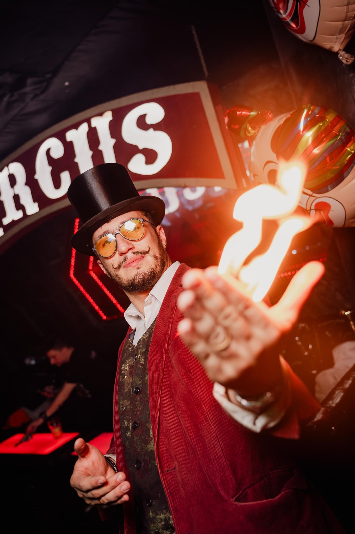 Uśmiechnięty iluzjonista w cylindrze i czerwonym fraku prezentuje płomień na dłoni, na tle dekoracji cyrkowych i balonów. Efektowne show z ogniem.