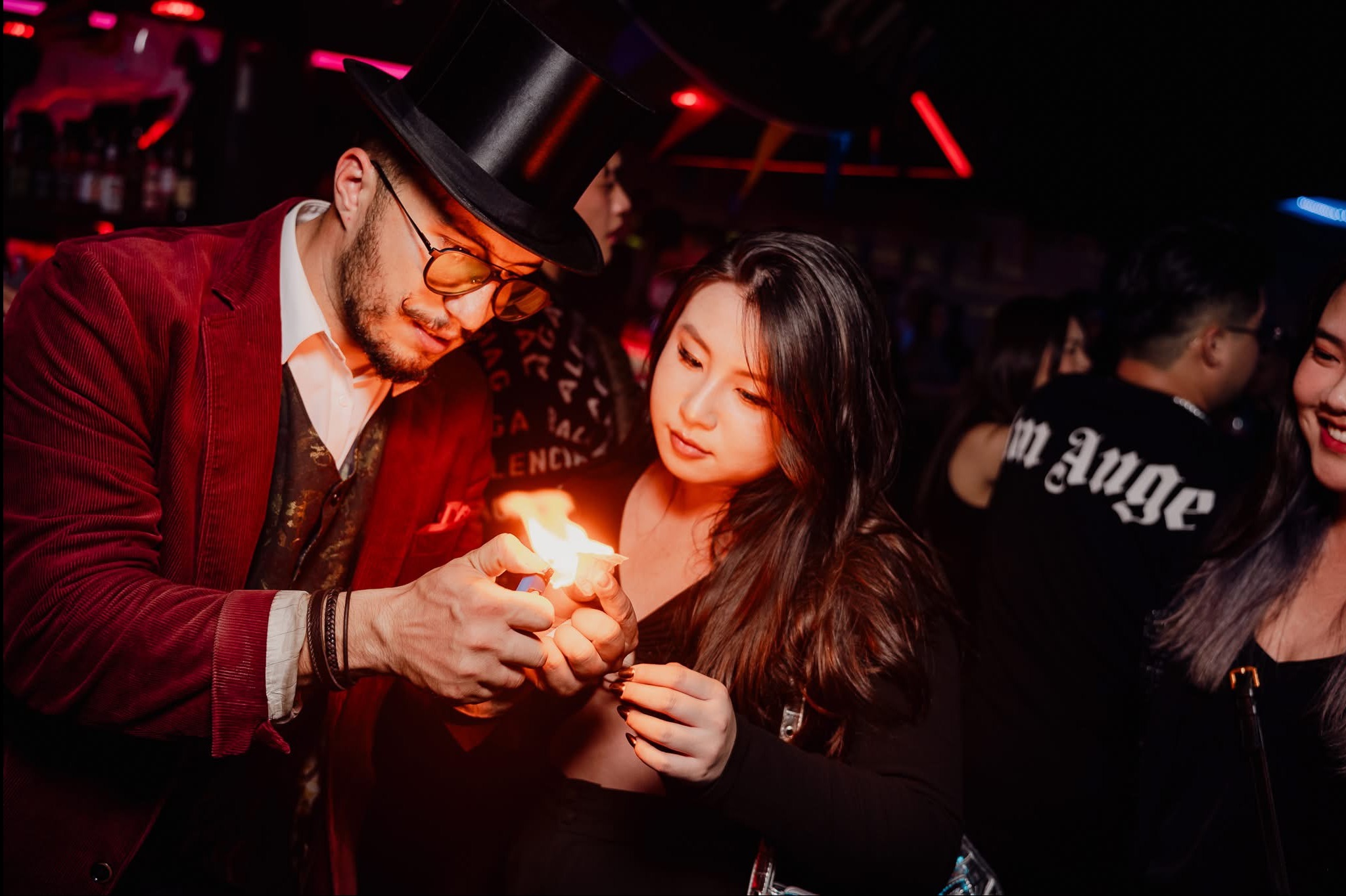 Iluzjonista w cylindrze podpala rekwizyt ogniem podczas występu dla publiczności w klubie. Czerwone światła i uśmiechnięte twarze w tle.