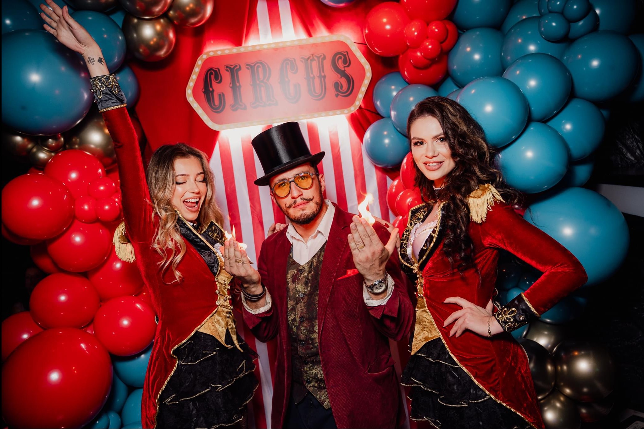 Uśmiechnięta grupa w kostiumach cyrkowych na tle czerwono-niebieskich balonów i dekoracji. Mężczyzna w cylindrze prezentuje płomienie w dłoniach.