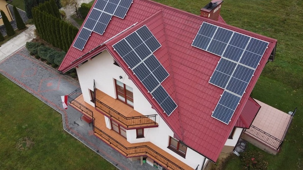 Widok z góry na dom jednorodzinny z czerwonym dachem i panelami słonecznymi, widoczny trawnik oraz chodnik z kostki.