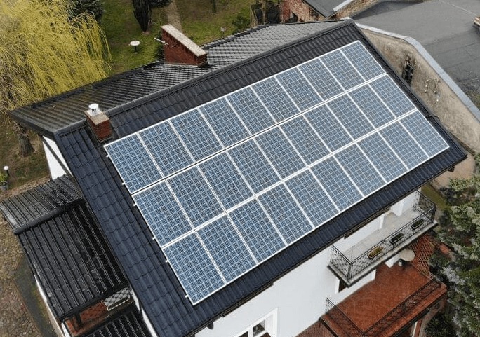 Instalacja paneli słonecznych na ciemnym dachu dwupiętrowego domu z balkonem, widok z góry.