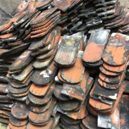 Stos starych, pomarańczowo-czarnych dachówek ceramicznych ułożonych warstwami przy szarej ścianie z cegieł, widoczne ślady użytkowania i zabrudzenia.