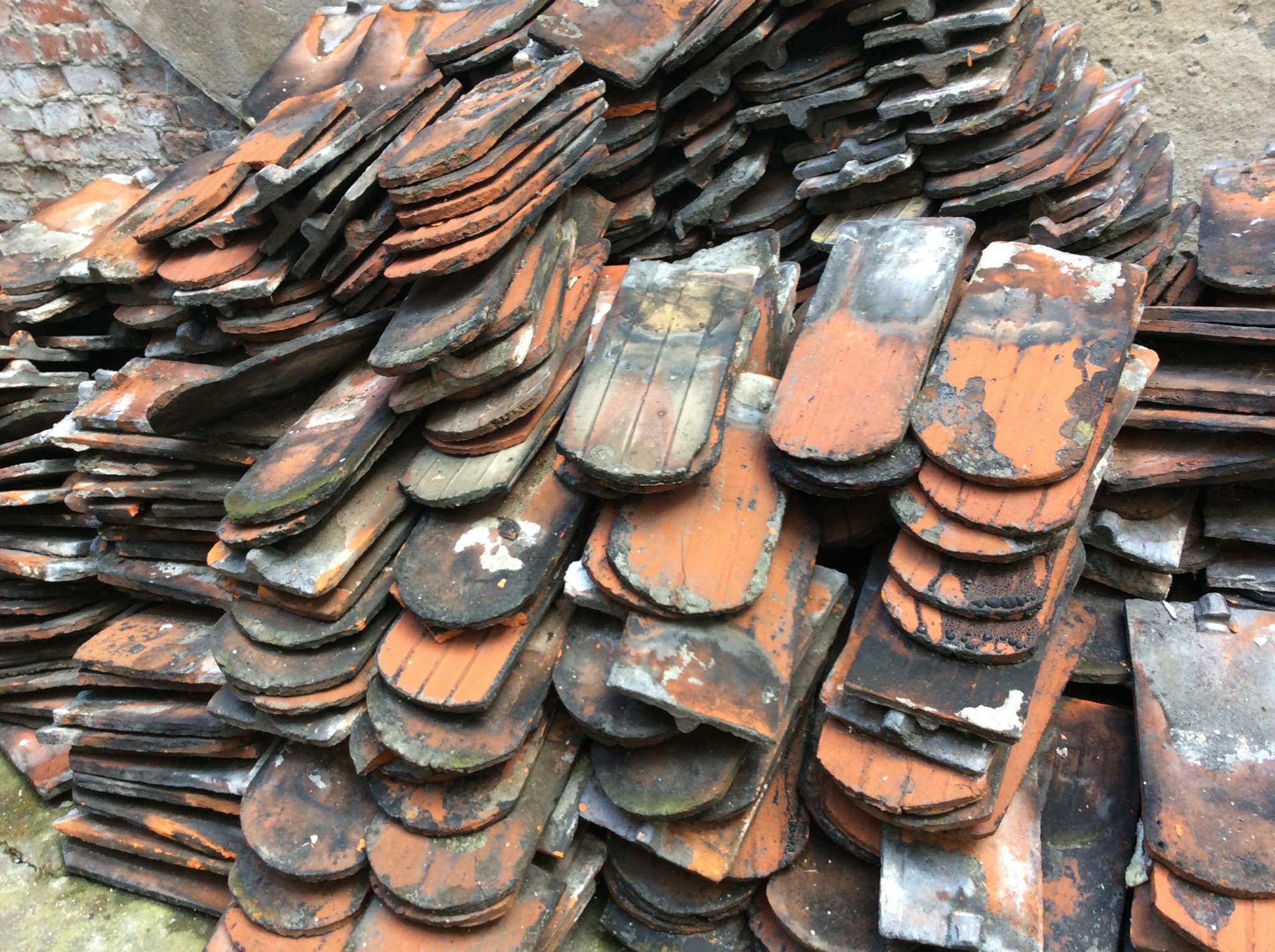 Stos starych, pomarańczowo-czarnych dachówek ceramicznych ułożonych warstwami przy szarej ścianie z cegieł, widoczne ślady użytkowania i zabrudzenia.