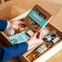 Otwarty drewniany box z prezentami: butelka wina Bukovino, konfitura jagodowa, pierniczki ręcznie robione w pudełku z napisem 'Pierniczki ręcznie robione', kartka z życzeniami 'Wesołych Świąt'...