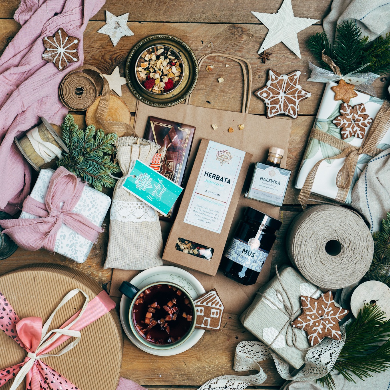 Kompozycja prezentowa na drewnianym stole z herbatą zimową, nalewką, dżemem, piernikami w kształcie gwiazdek i domków, zapakowanymi upominkami oraz dekoracjami świątecznymi z jutowego sznurka...
