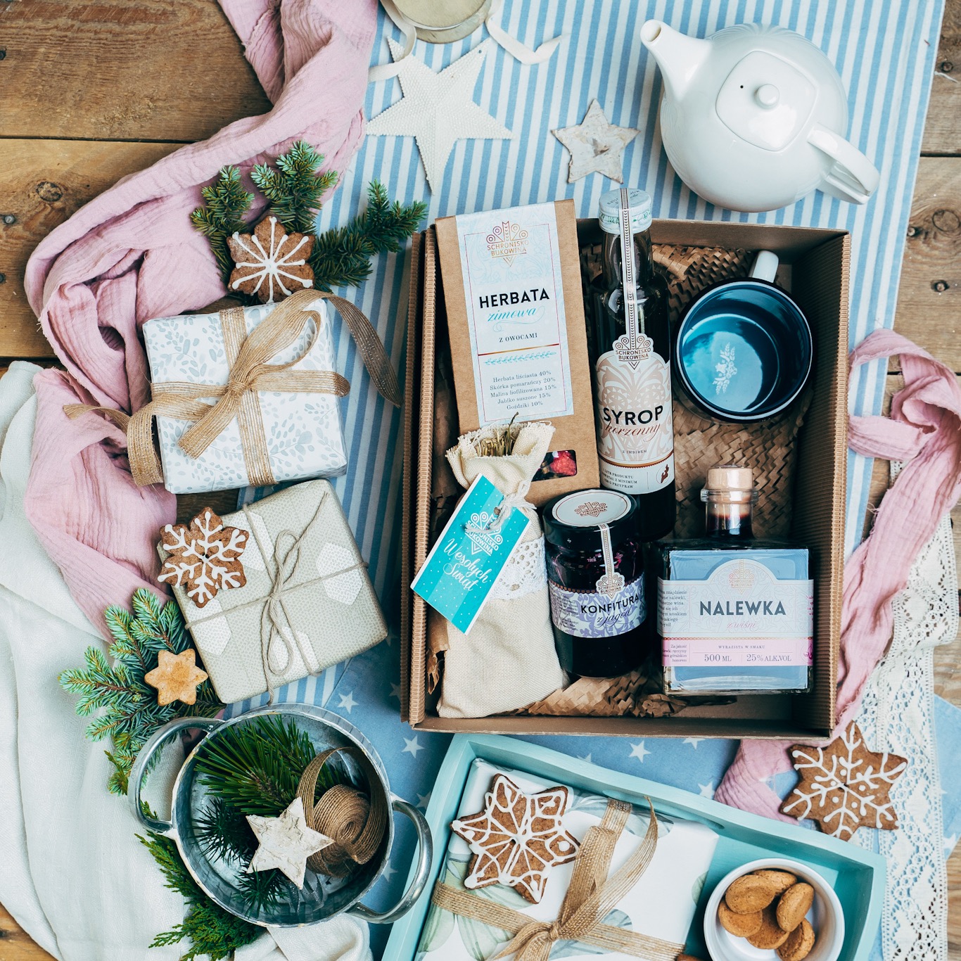 Świąteczny zestaw upominkowy z herbatą, syropem, konfiturą, nalewką, kubkiem, imbrykiem, ciastkami w kształcie gwiazdek, prezentami i dekoracjami na drewnianym stole z niebieskim bieżnikiem.
