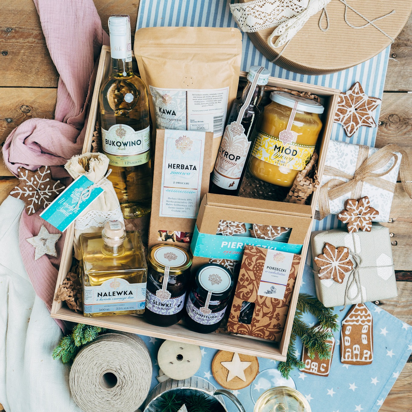 Drewniana skrzynka prezentowa na desce, zawierająca miód, kawę, herbatę, syrop, nalewkę, konfitury śliwkowe, czekoladę porzeczkową, pierniczki i butelkę wina Bukowino, uzupełniona ozdobami...