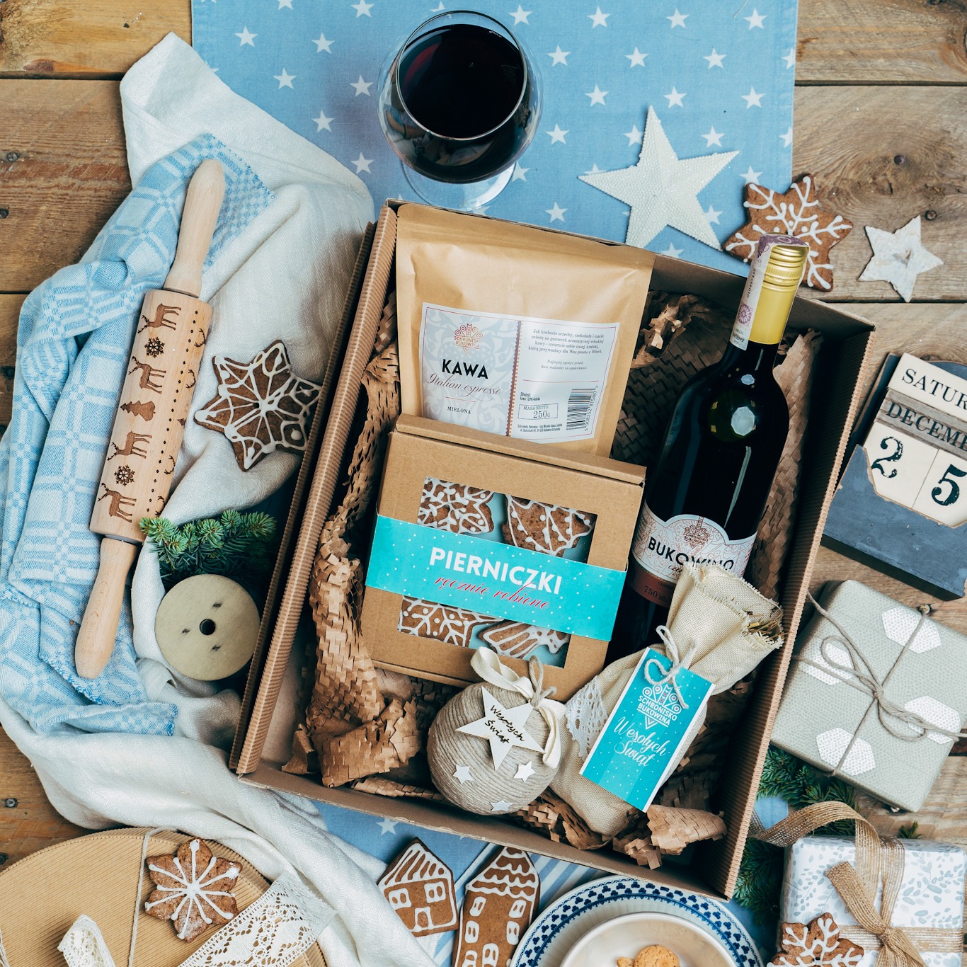 Świąteczny zestaw upominkowy: wino, kawa, pierniczki w pudełku z kalendarzem adwentowym, wałkiem do ciasta z reniferami, szklanką czerwonego wina i dekoracjami w kształcie gwiazd na drewnianym stole.