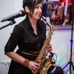 Saksofonistka w czarnej sukience i z kolorową bransoletką, grająca na złotym saksofonie na tle sceny z oświetleniem.