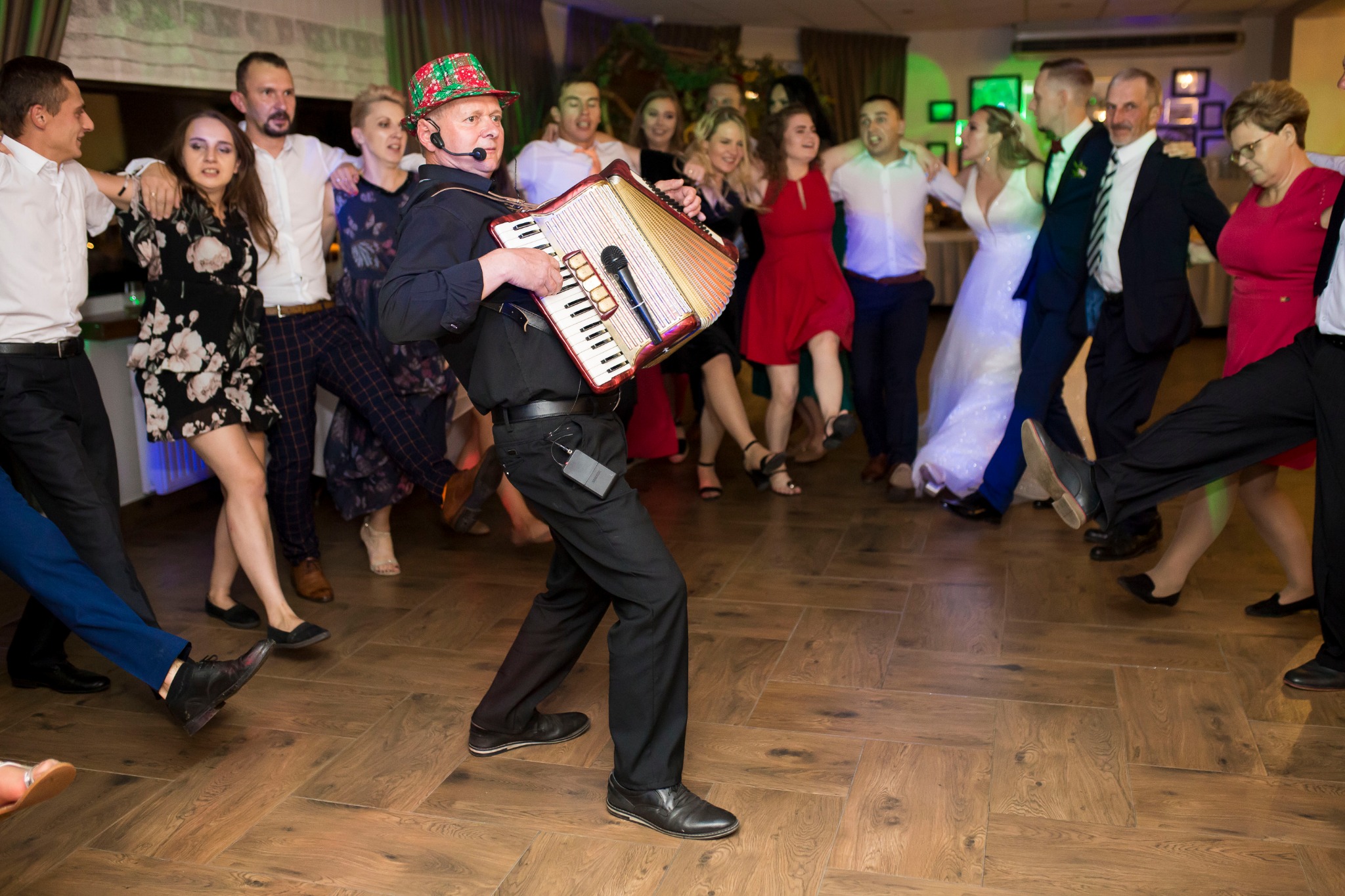 Akordeonista w kraciastej czapce gra podczas wesela, wokół tańczą goście w różnorodnych strojach, w tle widoczne dekoracje sali.