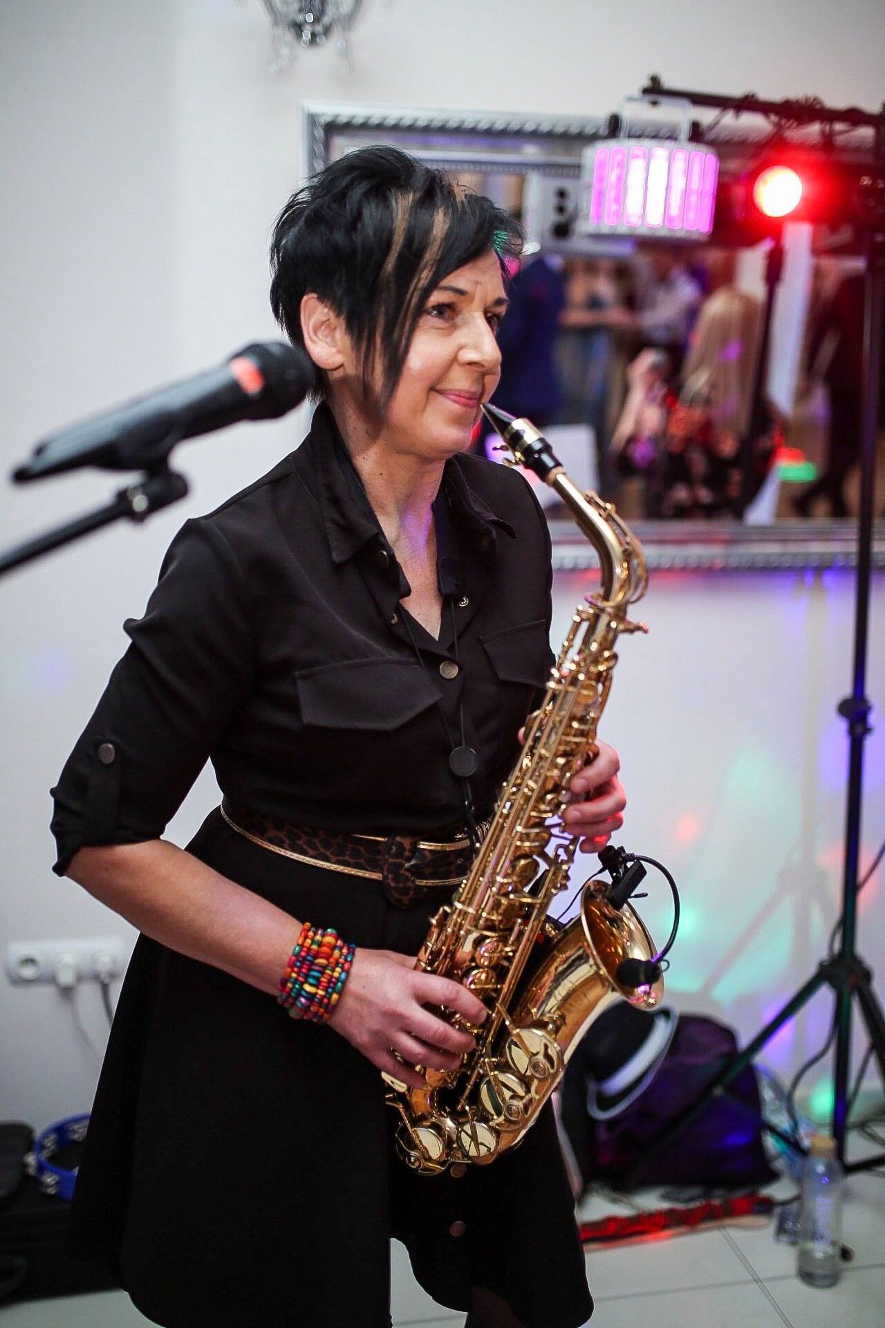 Saksofonistka w czarnej sukience i z kolorową bransoletką, grająca na złotym saksofonie na tle sceny z oświetleniem.