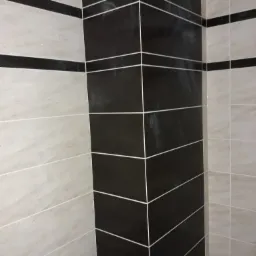 Fragment wykończonej łazienki z czarno-beżowymi płytkami ułożonymi w poziome pasy, widoczny fragment instalacji wodnej z czerwonym korkiem.