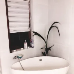 Nowoczesna biała wanna wolnostojąca w jasnej łazience z rośliną doniczkową i oknem z żaluzjami.