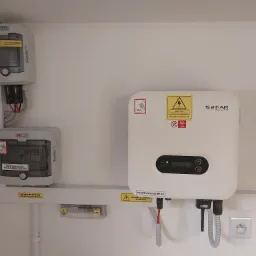 Biały inwerter Sofar Solar z podłączonymi kablami, obok rozdzielnica PV AC i licznik energii elektrycznej Noark, zamontowane na ścianie wewnątrz budynku.