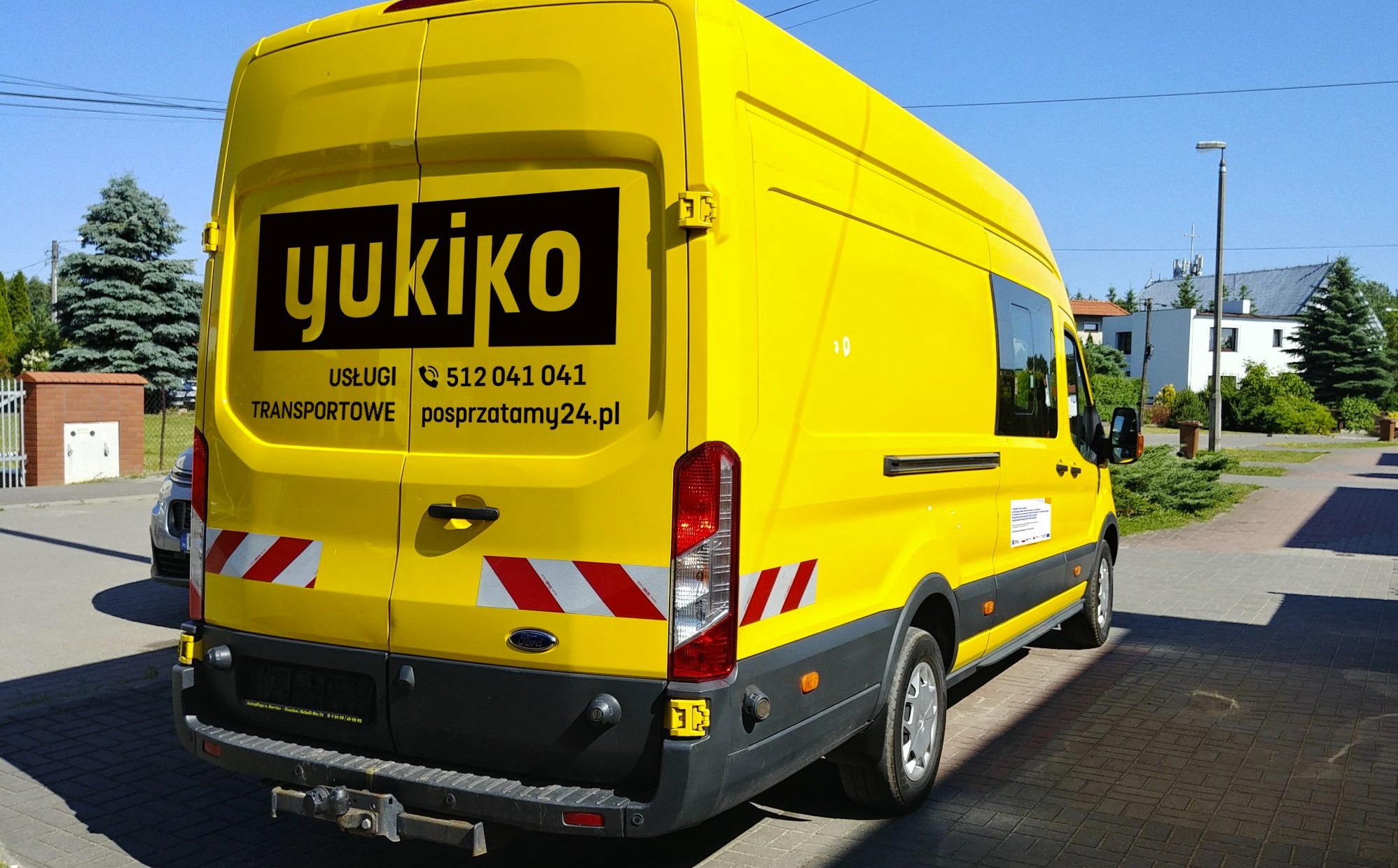 Żółty bus firmy Yukiko z Bydgoszczy, z widocznym logo i numerem telefonu, zaparkowany na chodniku przed budynkiem.