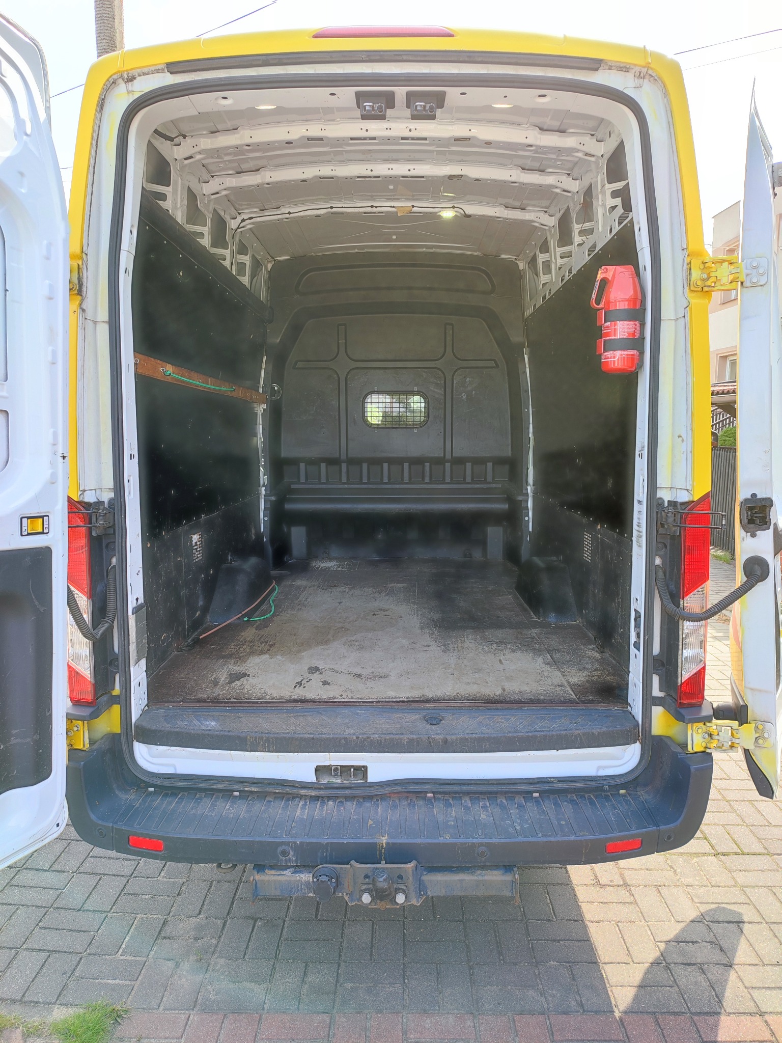 Wnętrze puste, brudne, białego busa z żółtymi elementami, widok z tyłu na otwarte drzwi, na ścianie gaśnica.