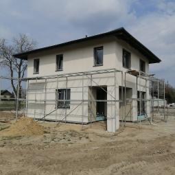 Dwukondygnacyjny dom w trakcie prac elewacyjnych, z widocznym rusztowaniem i ociepleniem ścian. Jasna elewacja, ciemny dach, błękitne niebo w tle.