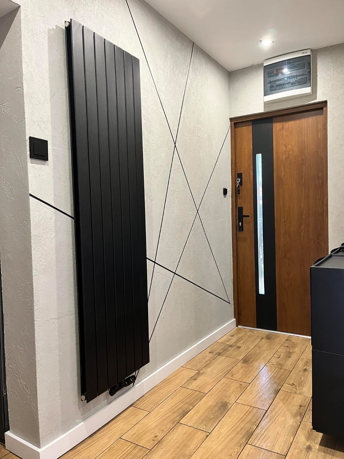 Nowoczesny przedpokój z czarnym grzejnikiem panelowym na ścianie z dekoracyjnymi liniami. Drewniane drzwi wejściowe i podłoga z paneli. Minimalistyczny design.