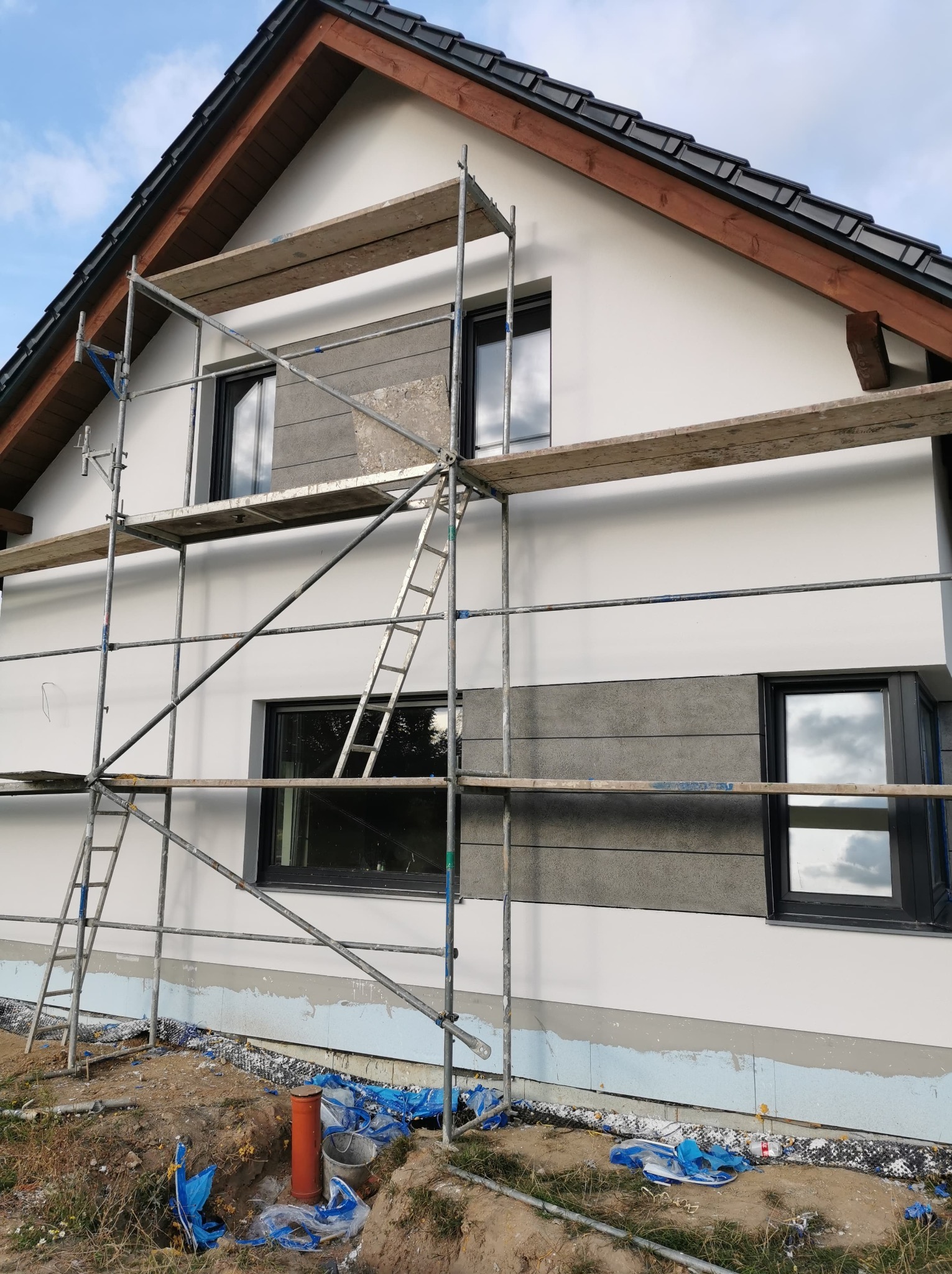 Dom jednorodzinny w trakcie ocieplania elewacji, widoczne rusztowania, fragmenty ocieplenia i wykończenia. Częściowo zakończone prace.