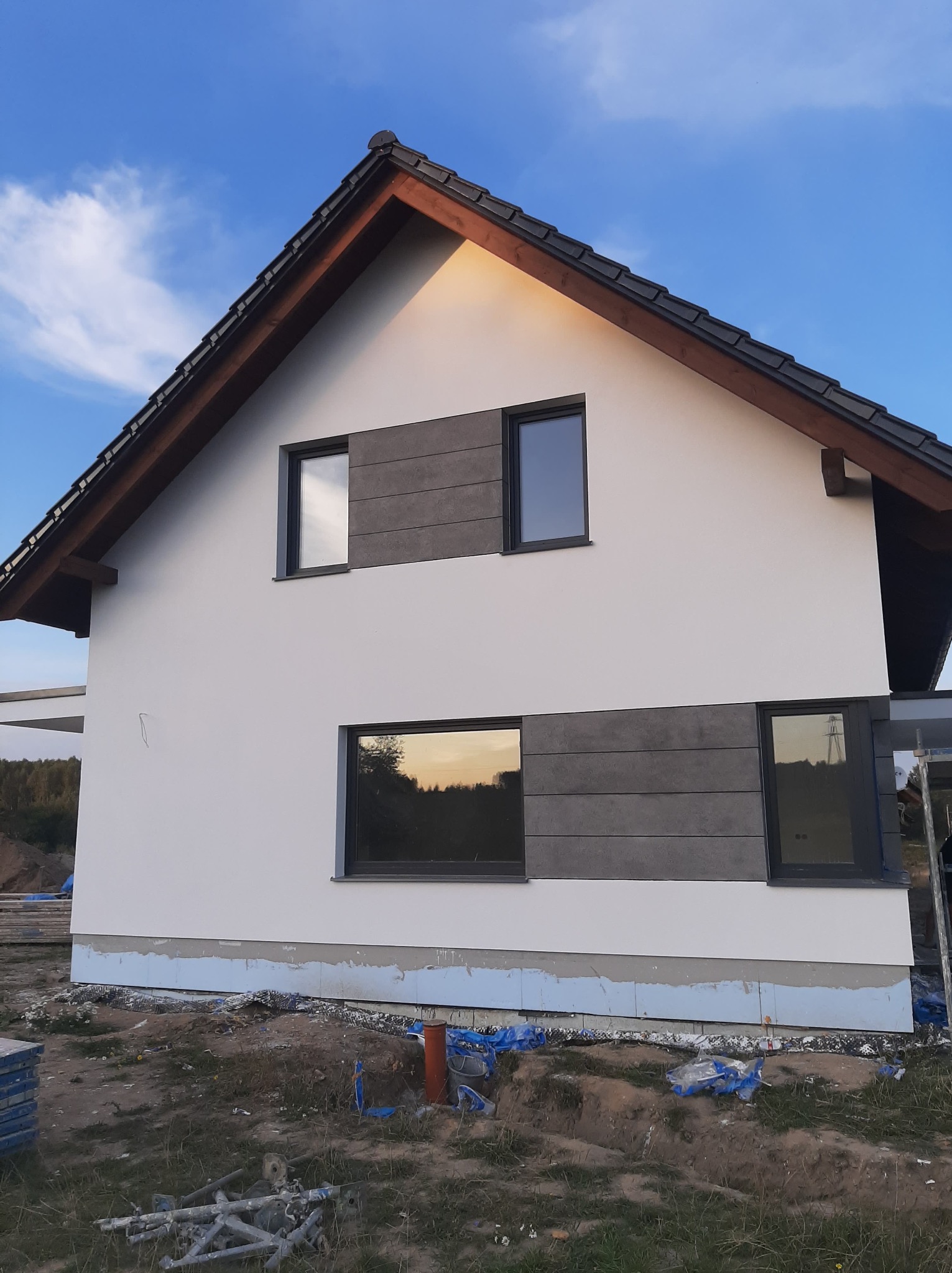 Boczny widok domu z białą elewacją i szarymi panelami, ciemnym dachem i oknami. Widoczne rusztowania i teren budowy. Okna odbijają zachód słońca.
