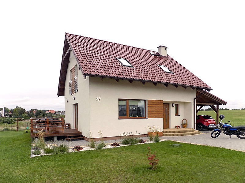 Jasny dom jednorodzinny z czerwoną dachówką, drewnianym tarasem i podjazdem. Widoczne elementy architektury krajobrazu i pojazdy.