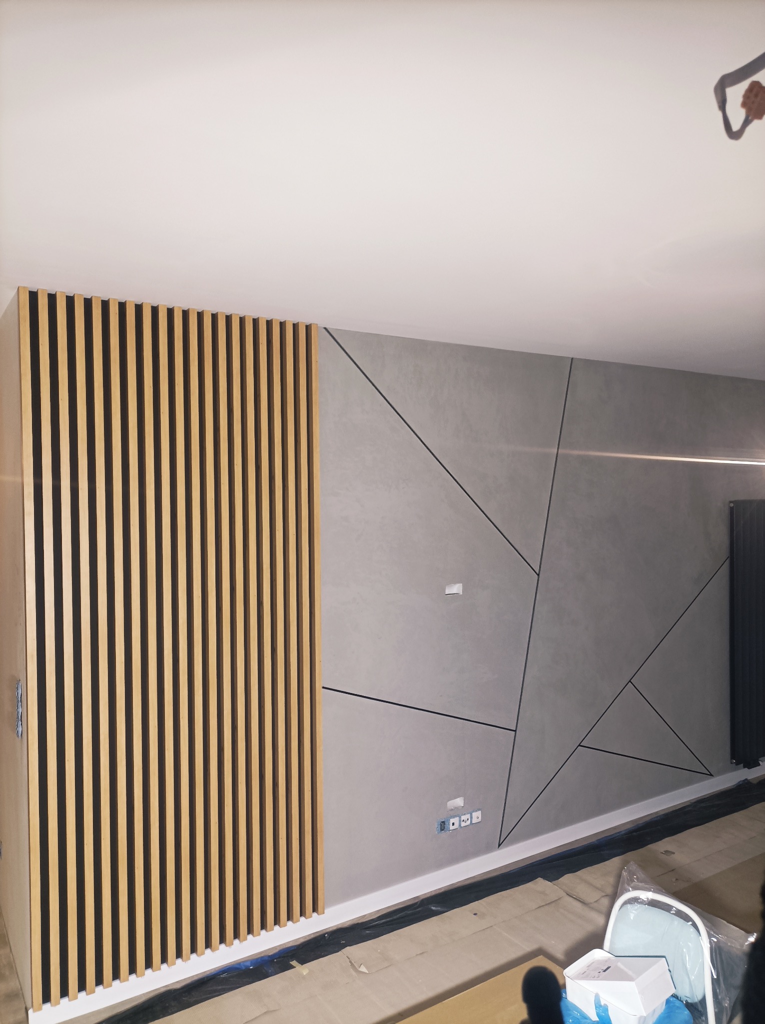 Nowoczesna ściana z dekoracyjną szarą gładzią i drewnianymi lamelami. Geometryczny wzór z czarnymi liniami. Widoczne gniazdka elektryczne i fragment grzejnika.