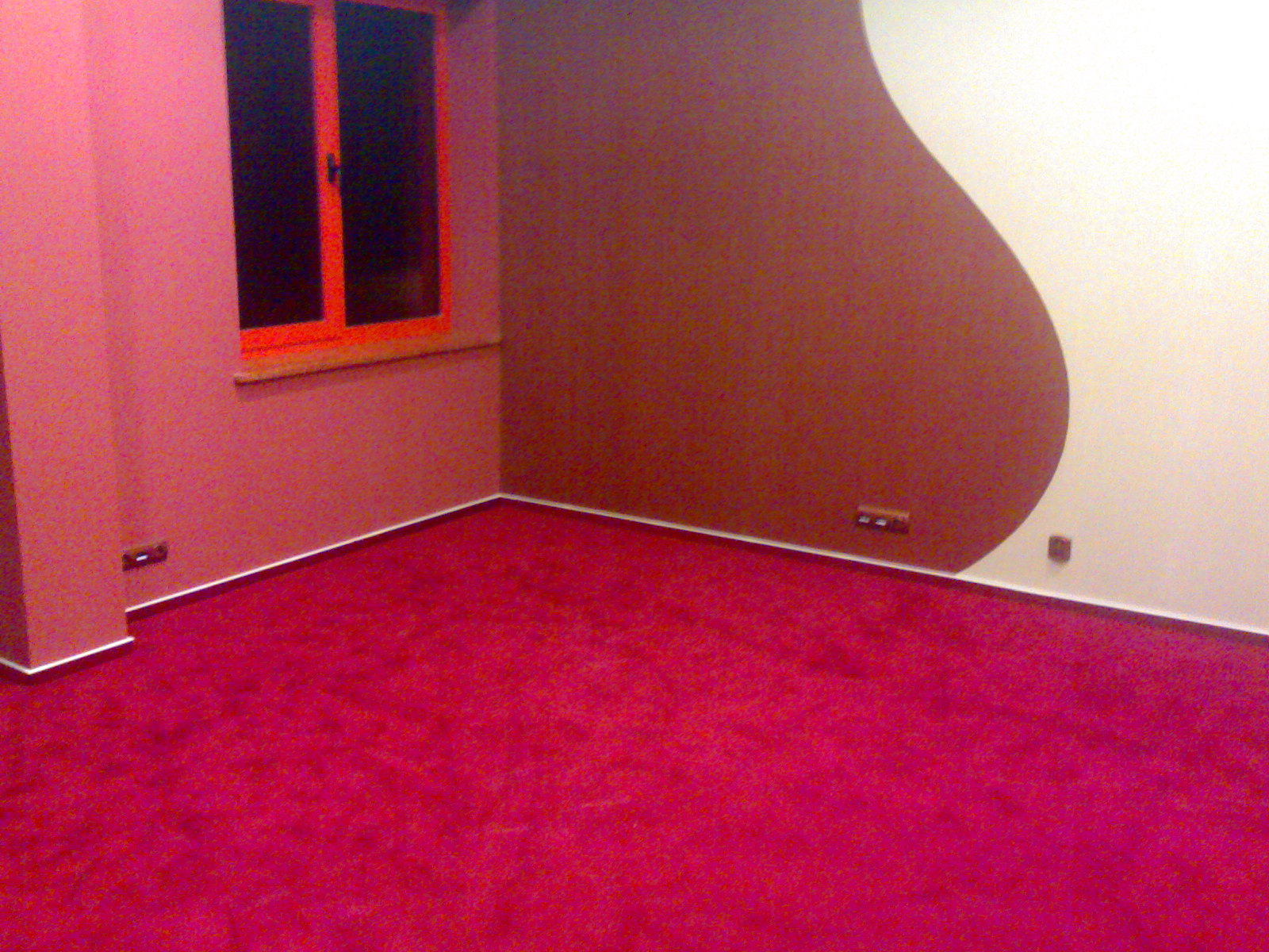 Pokój z intensywnie różową wykładziną dywanową, ścianami w odcieniach różu i brązu oraz pomarańczową ramą okienną. Dekoracyjny, brązowy element na ścianie. Widoczne gniazdka elektryczne.