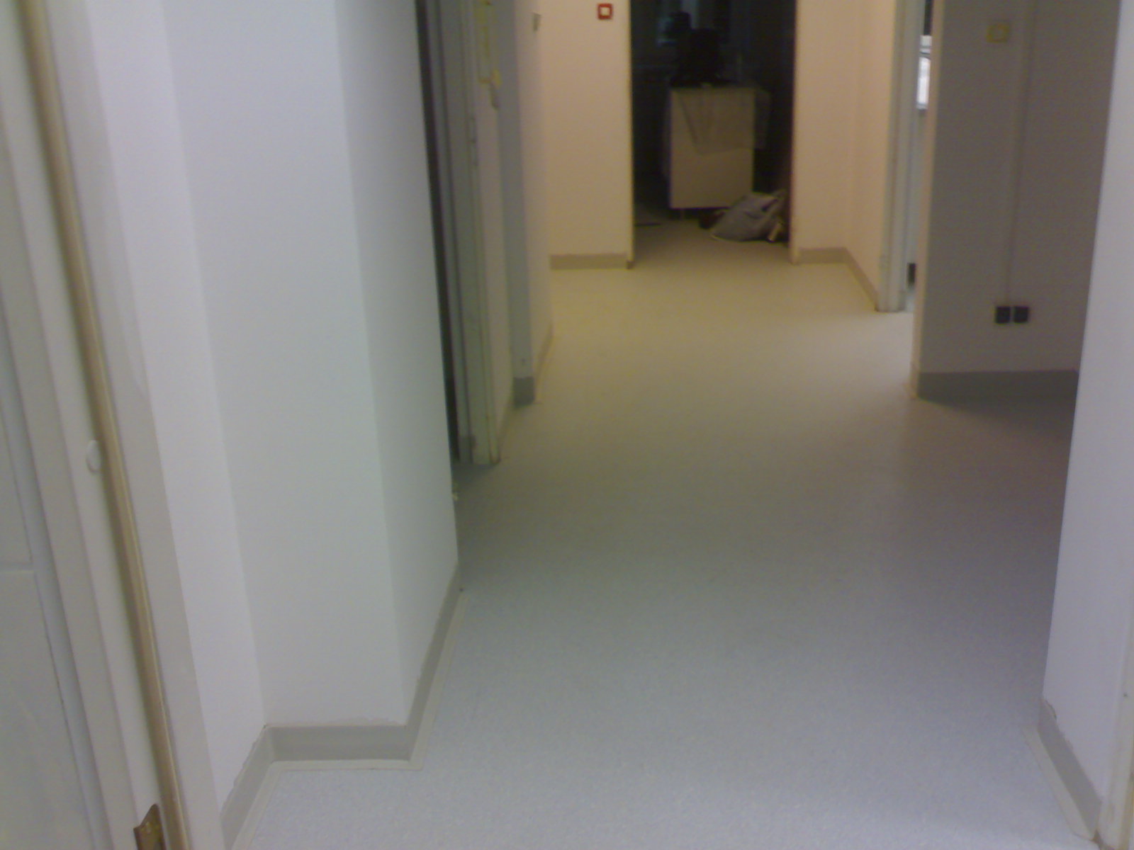 Korytarz z nową, jasną wykładziną PCV, widoczne białe ściany i listwy przypodłogowe, perspektywa zza otwartych drzwi.