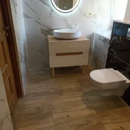 Nowoczesna łazienka z drewnopodobną podłogą, marmurowymi płytkami ściennymi, okrągłym lustrem z podświetleniem LED oraz białą umywalką nablatową na szafce z drewnianymi nogami i wiszącą toaletą.