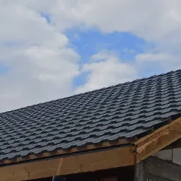 Fragment dachu z czarnej blachodachówki trapezowej, widoczne krokwie i fragment ściany z pustaków, na tle błękitnego nieba z białymi chmurami.