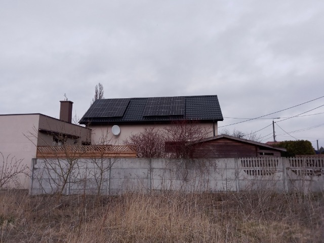 Dom jednorodzinny z panelami słonecznymi na dachu, widoczne zza betonowego ogrodzenia i uschniętej trawy w pochmurny dzień.