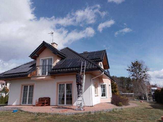 Montaż paneli słonecznych na dachu domu jednorodzinnego; pracownik na drabinie mocuje panele, widoczne jasne niebo z chmurami.