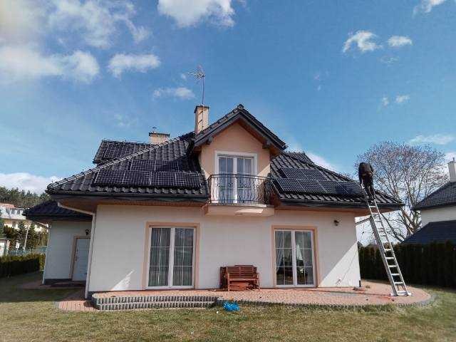 Dom jednorodzinny z zamontowanymi panelami słonecznymi na dachu; pracownik na drabinie wykonuje prace serwisowe.