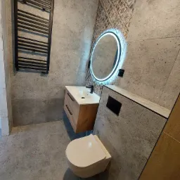 Nowoczesna łazienka z szarymi płytkami imitującymi kamień, podświetlanym okrągłym lustrem, wiszącą szafką z umywalką i grzejnikiem drabinkowym w kolorze czarnym.