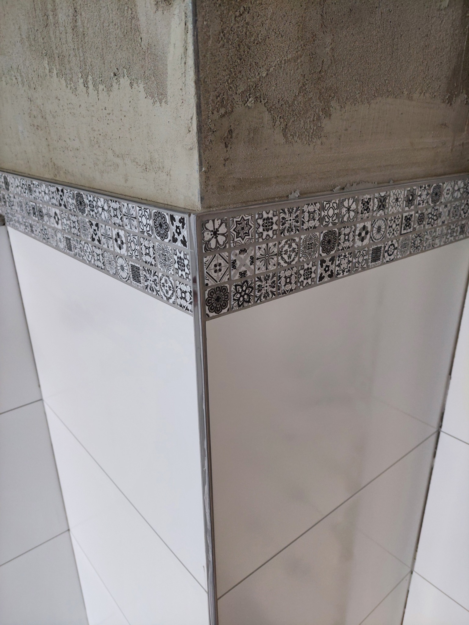 Wykończony narożnik ściany z białymi płytkami i dekoracyjnym pasem mozaiki z motywami kwiatowymi i geometrycznymi, widoczny surowy beton powyżej.