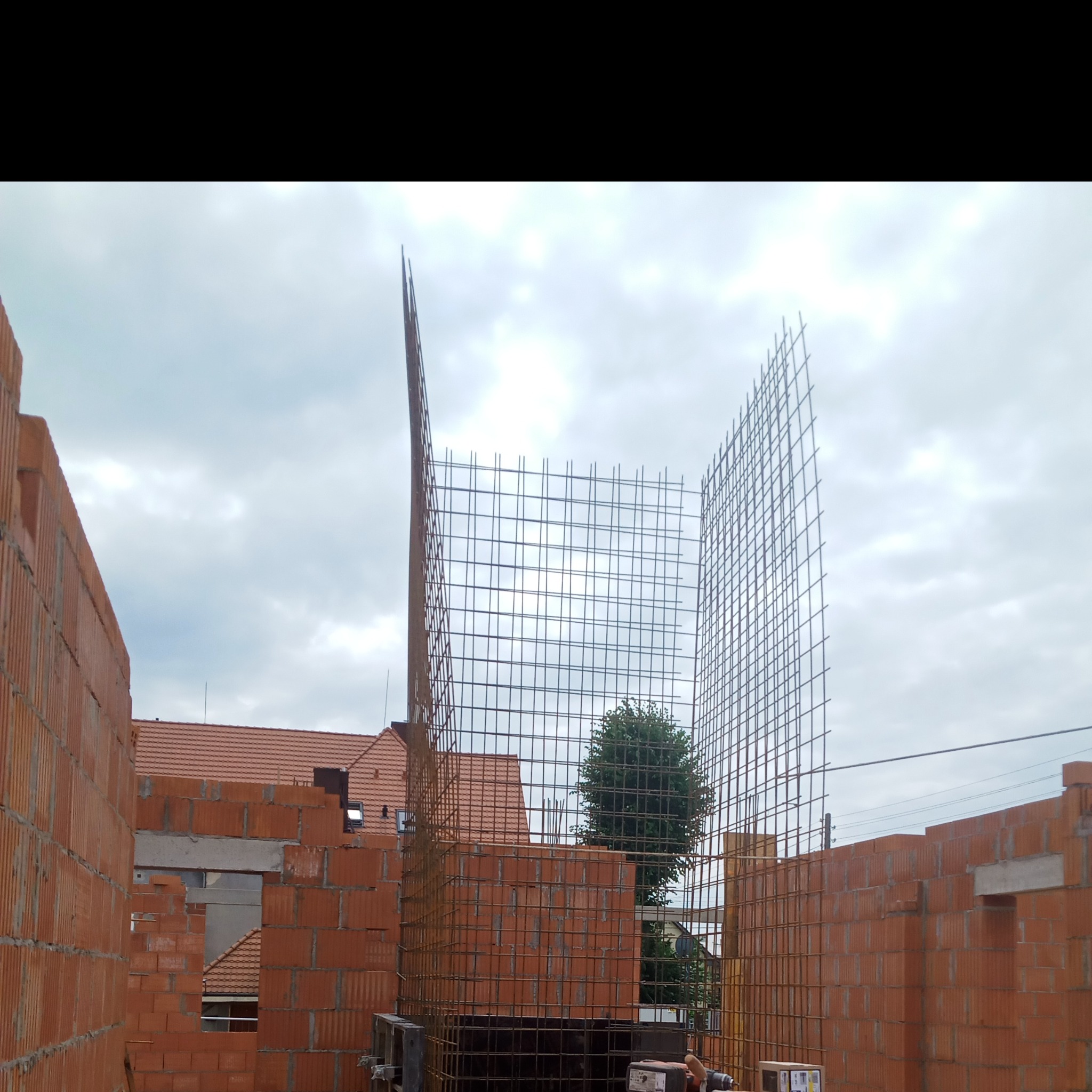 Wznoszone ściany z czerwonej cegły z widocznym zbrojeniem na budowie domu jednorodzinnego, na tle pochmurnego nieba.