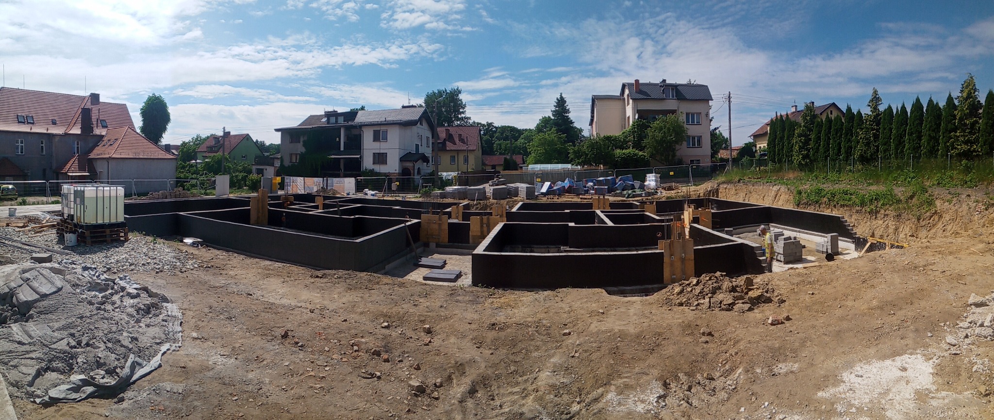 Szeroki kadr na wykop z izolowanymi fundamentami domu jednorodzinnego, widoczne szalunki i bloczki betonowe, w tle zabudowa mieszkalna i błękitne niebo z chmurami.